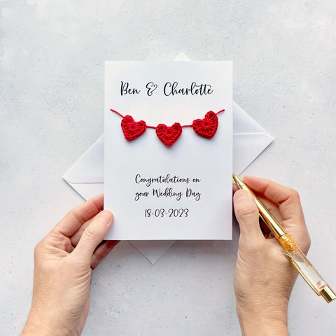 A white card with a string of 3 red crochet hearts across the front. Above the hearts are the two names and at the bottom of the card is 'Congratulations on your wedding day' and the wedding date. 