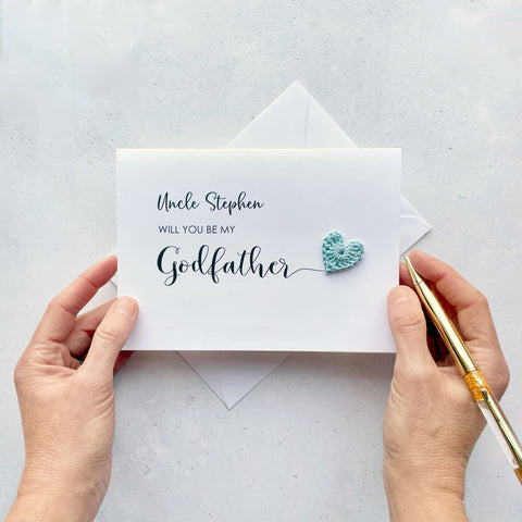 A white card with black cursive text printed on it. It reads 'Uncle Stephen, Will you be my Godfather’. The word ‘Godfather’ has been printed larger than the rest and it has a little pale blue crochet heart attatched to the ‘r’.