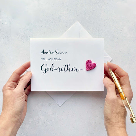 A white card with black cursive text printed on it. It reads ‘Auntie Emma, Will you be my Godmother’. The word ‘Godmother’ has been printed larger than the rest and it has a little bright pink crochet heart attatched to the ‘r’.