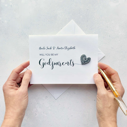 A white card with black cursive text printed on it. It reads 'Uncle Jack & Auntie Elizabeth, Will you be my Godparents'. The word 'godparents'has been printed larger than the rest and it has a little grey crochet heart attatched to the 's'.