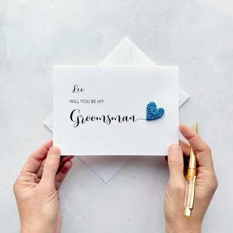A white card with ‘Will you be my Groomsman’ printed on it. The word ‘Groomsman’ appears larger than the rest of the text and it has a little blue crochet heart at the end of the word.  The name ‘Lee’ has been printed at the top of the text, making this card a lovely keepsake. 