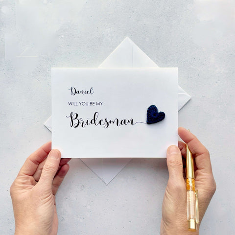 A white card with ‘Will you be my Bridesman’ printed on it. The word ‘Bridesman’ appears larger than the rest of the text and it has a little navy blue crochet heart at the end of the word.  The name ‘Daniel’ has been printed at the top of the text, making this card a lovely keepsake. 