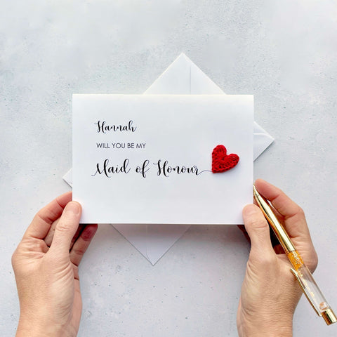 A white card with ‘Will you be my Maid of Honour’ printed on it. The words ‘Maid of Honour’ appears larger than the rest of the text and it has a little red crochet heart at the end of the word.  The name ‘Hannah’ has been printed at the top of the text, making this card a lovely keepsake. 