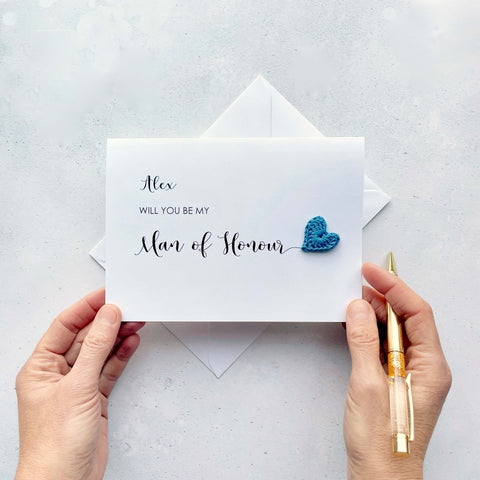 A white card with ‘Will you be my Man of Honour’ printed on it. The words ‘Man of Honour’ appears larger than the rest of the text and it has a little blue crochet heart at the end of the word.  The name ‘Alex’ has been printed at the top of the text, making this card a lovely keepsake. 