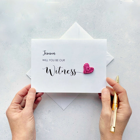 A white card with ‘Will you be our Witness’ printed on it. The word ‘Witness’ appears larger than the rest of the text and it has a little bright pink crochet heart at the end of the word.  The name ‘Jemma’ has been printed at the top of the text, making this card a lovely keepsake. 