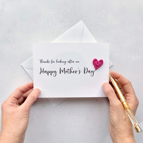 A white Mother’s Day card that has ‘Thanks for looking after me, Happy Mother’s Day’ printed on the card in black cursive text. A bright pink crochet heart has been attatched to the ‘y’ at the end. 