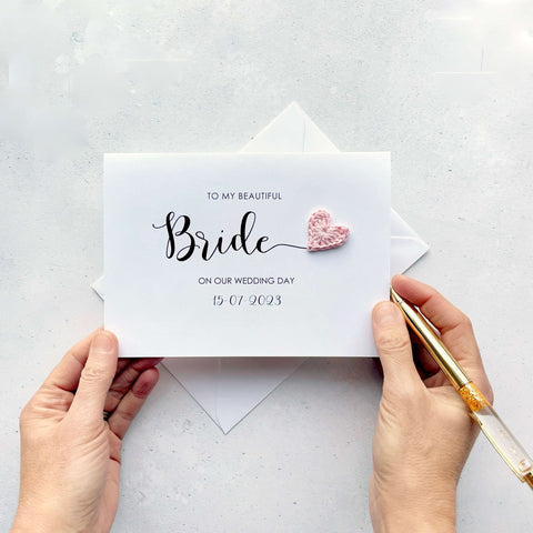 A white card with 'To my beautiful Bride on our wedding day' printed on the front of the card along with the wedding date. The word 'Bride' appears much larger than the rest and there is a pale pink crochet heart at the end of the letter 'e'