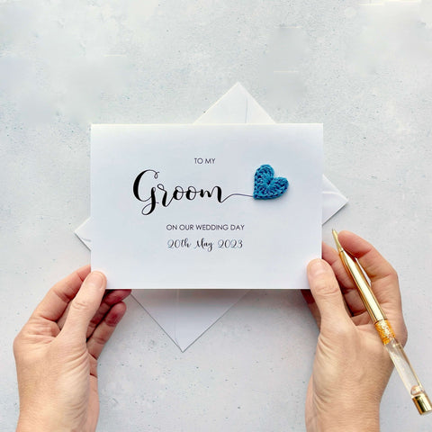 A white card with 'To my Groom on our wedding day' printed on the front. The wedding date is also printed on there. The word 'Groom' is much larger than the rest of the text and there is a blue crochet heart attached to the 'm' at the end. 