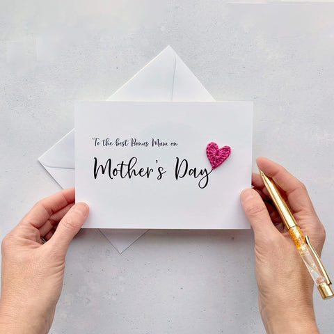 A white Mother’s Day card that has ‘To the best Bonus Mum on Mother’s Day’ printed on the card in black cursive text. A bright pink crochet heart has been attatched to the ‘y’ at the end. 