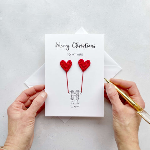 A white Christmas card featuring a line drawn couple holding hands and each holding a red crochet heart balloon. ‘Merry Christmas to my Wife’ is printed at the top of the card. The couple are drawn wearing winter scarves and santa hats.