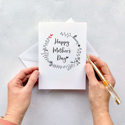 A white Mother’s Day card. Black line drawn foliage is printed on teh card in a circle pattern. The words ‘Happy Mother’s Day’ has been printed inside the circle in black cursive text. 