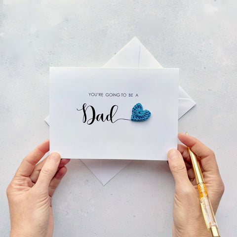 A white card with ‘You’re going to be a Dad’ printed on it. The word ‘Dad’ appears larger than the rest of the text and it has a little blue crochet heart at the end of the word.  Under the word ’Dad’ is the babies surname and due date making this card a lovely keepsake. 
