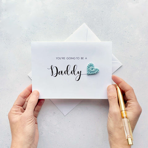 A white card with ‘You’re going to be a Daddy’ printed on it. The word ‘Daddy’ appears larger than the rest of the text and it has a little pale blue crochet heart at the end of the word. 