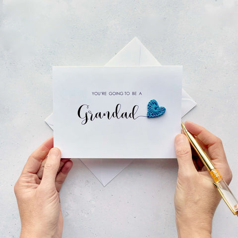 A white card with ‘You’re going to be Grandad’ printed on it. The word ‘Grandad’ appears larger than the rest of the text and it has a little blue crochet heart at the end of the word.  