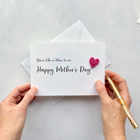 A white Mother’s Day card that has ‘You’re like a Mum to me , Happy Mother’s Day’ printed on the card in black cursive text. A bright pink crochet heart has been attatched to the ‘y’ at the end. 