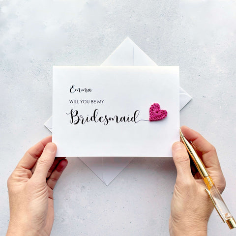 A white card with ‘Will you be my Bridesmaid’ printed on it. The word ‘Bridesmaid’ appears larger than the rest of the text and it has a little bright pink crochet heart at the end of the word.  The name ‘Emma’ has been printed at the top of the text, making this card a lovely keepsake. 