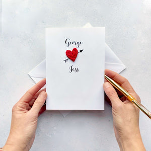 A Valentines Day greeting card featuring a red crochet heart with a black cupid arrow through the back of it. The name George is printed in black above the heart and Jess printed below the heart. 