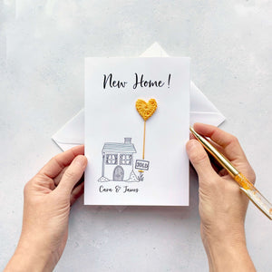 A new Home card on white card. Black cursive script text appears at the top reading ‘New Home’. There is a simple black line drawn house on the front of the card and there’s a pale blue cotton crochet heart balloon coming up from the 'sold' sign. 