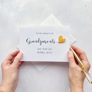 A white personalised pregnancy reveal card with black cursive script text. The text reads ‘You’re going to be Grandparents, baby White due October 2023’ finished with a mini yellow crochet heart.