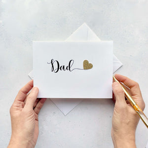 A white card with the word 'Dad' on the front  - the text is black cursive script. There is a sparkly gold heart stuck on the front of the card too. 