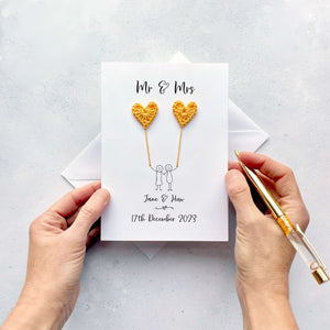 A personalised wedding card with black cursive script text on white card. The card reads 'Mr & Mrs' at the top and 'James and Katie' at the bottom. The main focus on the card is a line drawn couple each holding a mini yellow crochet heart.