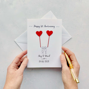 A paper wedding anniversary card featuring a line drawn couple each holding a heart shaped balloon made from red card. ’Happy 1st Anniversary' is printed at the top of the card and the couples names and wedding date are printed at the bottom. 