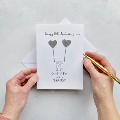 The image shows a beautiful handmade anniversary card with two hearts made from steel coloured card, these are attached by grey yarn, resembling balloons being held by a hand-drawn couple underneath. The text on the card reads "Happy 11th Anniversary" at the top, followed by the couples names and wedding date at the bottom. The card is set on a light background, and the person's hands are seen holding a pen and gently holding the card, giving the image a sense of care and personalisation.