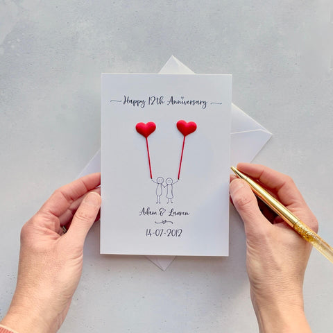 The image shows a beautiful handmade anniversary card with small red silk padded hearts, these are attached by red yarn, resembling balloons being held by a hand-drawn couple underneath. The text on the card reads "Happy 12th Anniversary" at the top, followed by the couples names and wedding date at the bottom. The card is set on a light background, and the person's hands are seen holding a pen and gently holding the card, giving the image a sense of care and personalisation.