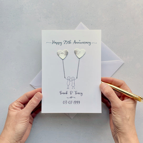 The image shows a beautiful handmade anniversary card with mirror silver card hearts, these are attached by cotton yarn, resembling balloons being held by a hand-drawn couple underneath. The text on the card reads "Happy 25th Anniversary" at the top, followed by the couples names and wedding date at the bottom. The card is set on a light background, and the person's hands are seen holding a pen and gently holding the card, giving the image a sense of care and personalisation.