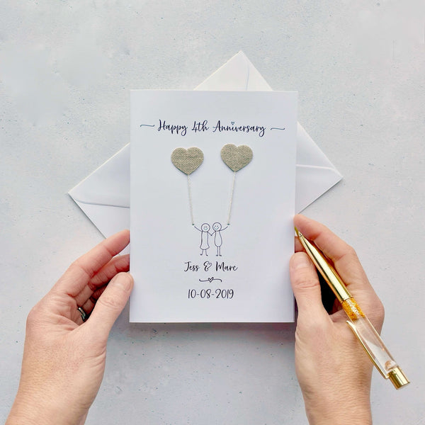 A white card featuring a line drawn couple each holding a heart shaped balloon which is made from linen. Happy 4th Anniversary is printed at the top of the card and the couples names and wedding date are printed at the bottom. 