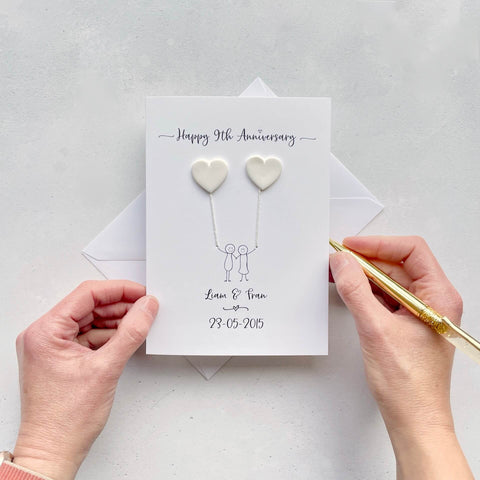 The image shows a beautiful handmade anniversary card with two hearts made from white polymer clay, these are attached by ivory yarn, resembling balloons being held by a hand-drawn couple underneath. The text on the card reads "Happy 9th Anniversary" at the top, followed by the couples names and wedding date at the bottom. The card is set on a light background, and the person's hands are seen holding a pen and gently holding the card, giving the image a sense of care and personalisation.