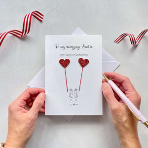 A Christmas card featuring a minimalist design with two figures wearing santa hats holding red glitter heart balloons. The text at the top reads: "To my amazing Auntie, With Love at Christmas." The card is placed on a white background with a white envelope partially visible behind it. A hand is seen holding the card, while another hand holds a pink pen. A red and white striped ribbon is positioned in the top corners of the image for decoration.