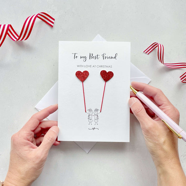 A Christmas card featuring a minimalist design with two figures wearing santa hats holding red glitter heart balloons. The text at the top reads: ‘To my Best Friend, withh love at Christmas’. The card is placed on a white background with a white envelope partially visible behind it. A hand is seen holding the card, while another hand holds a pink pen. A red and white striped ribbon is positioned in the top corners of the image for decoration.