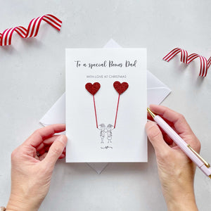 A Christmas card featuring a minimalist design with two figures wearing santa hats holding red glitter heart balloons. The text at the top reads: ‘To a special Bonus Dad, with love at Christmas’. The card is placed on a white background with a white envelope partially visible behind it. A hand is seen holding the card, while another hand holds a pink pen. A red and white striped ribbon is positioned in the top corners of the image for decoration.