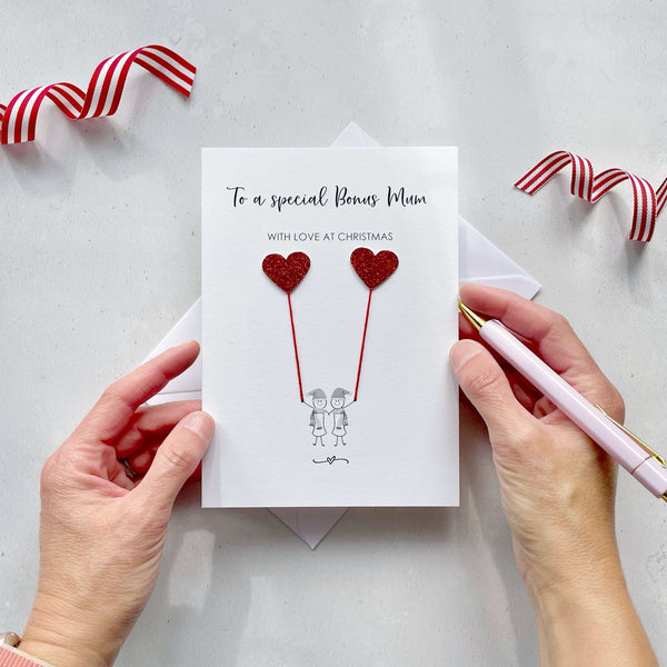 A Christmas card featuring a minimalist design with two figures wearing santa hats holding red glitter heart balloons. The text at the top reads: ‘To a special Bonus Mum, with love at Christmas’. The card is placed on a white background with a white envelope partially visible behind it. A hand is seen holding the card, while another hand holds a pink pen. A red and white striped ribbon is positioned in the top corners of the image for decoration.