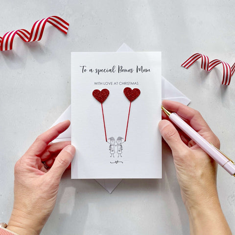 A Christmas card featuring a minimalist design with two figures wearing santa hats holding red glitter heart balloons. The text at the top reads: ‘To a special Bonus Mum, with love at Christmas’. The card is placed on a white background with a white envelope partially visible behind it. A hand is seen holding the card, while another hand holds a pink pen. A red and white striped ribbon is positioned in the top corners of the image for decoration.