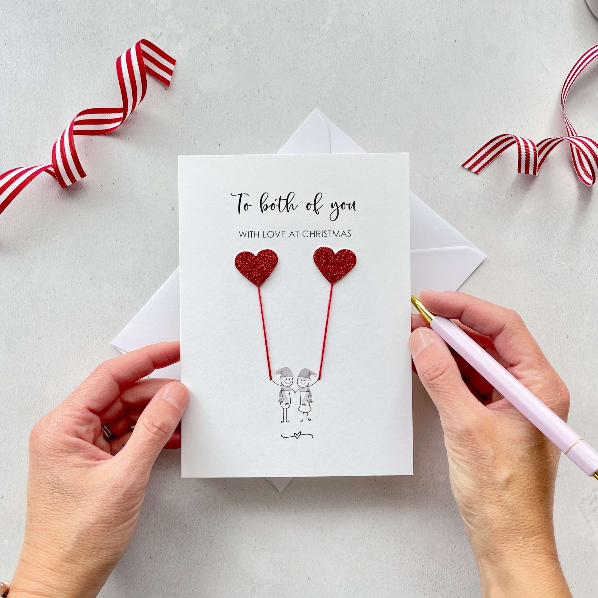 A Christmas card featuring a minimalist design with two figures wearing santa hats holding red glitter heart balloons. The text at the top reads: ‘To both of you, with love at Christmas’. The card is placed on a white background with a white envelope partially visible behind it. A hand is seen holding the card, while another hand holds a pink pen. A red and white striped ribbon is positioned in the top corners of the image for decoration.