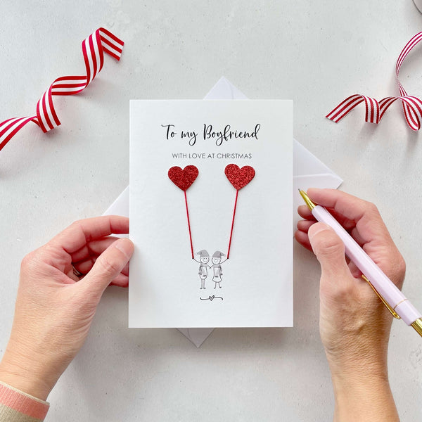 A Christmas card featuring a minimalist design with two figures wearing santa hats holding red glitter heart balloons. The text at the top reads: ‘To my Boyfriend, with love at Christmas’. The card is placed on a white background with a white envelope partially visible behind it. A hand is seen holding the card, while another hand holds a pink pen. A red and white striped ribbon is positioned in the top corners of the image for decoration.