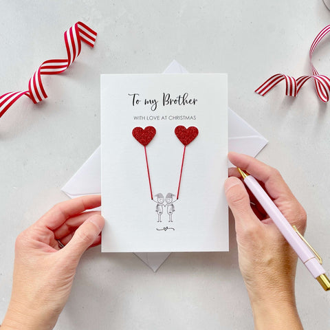 A Christmas card featuring a minimalist design with two figures wearing santa hats holding red glitter heart balloons. The text at the top reads: ‘To my Brother, With Love at Christmas’. The card is placed on a white background with a white envelope partially visible behind it. A hand is seen holding the card, while another hand holds a pink pen. A red and white striped ribbon is positioned in the top corners of the image for decoration.