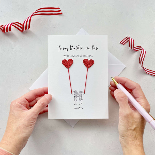 A Christmas card featuring a minimalist design with two figures wearing santa hats holding red glitter heart balloons. The text at the top reads: ‘To my Brother in law, with love at Christmas’. The card is placed on a white background with a white envelope partially visible behind it. A hand is seen holding the card, while another hand holds a pink pen. A red and white striped ribbon is positioned in the top corners of the image for decoration.