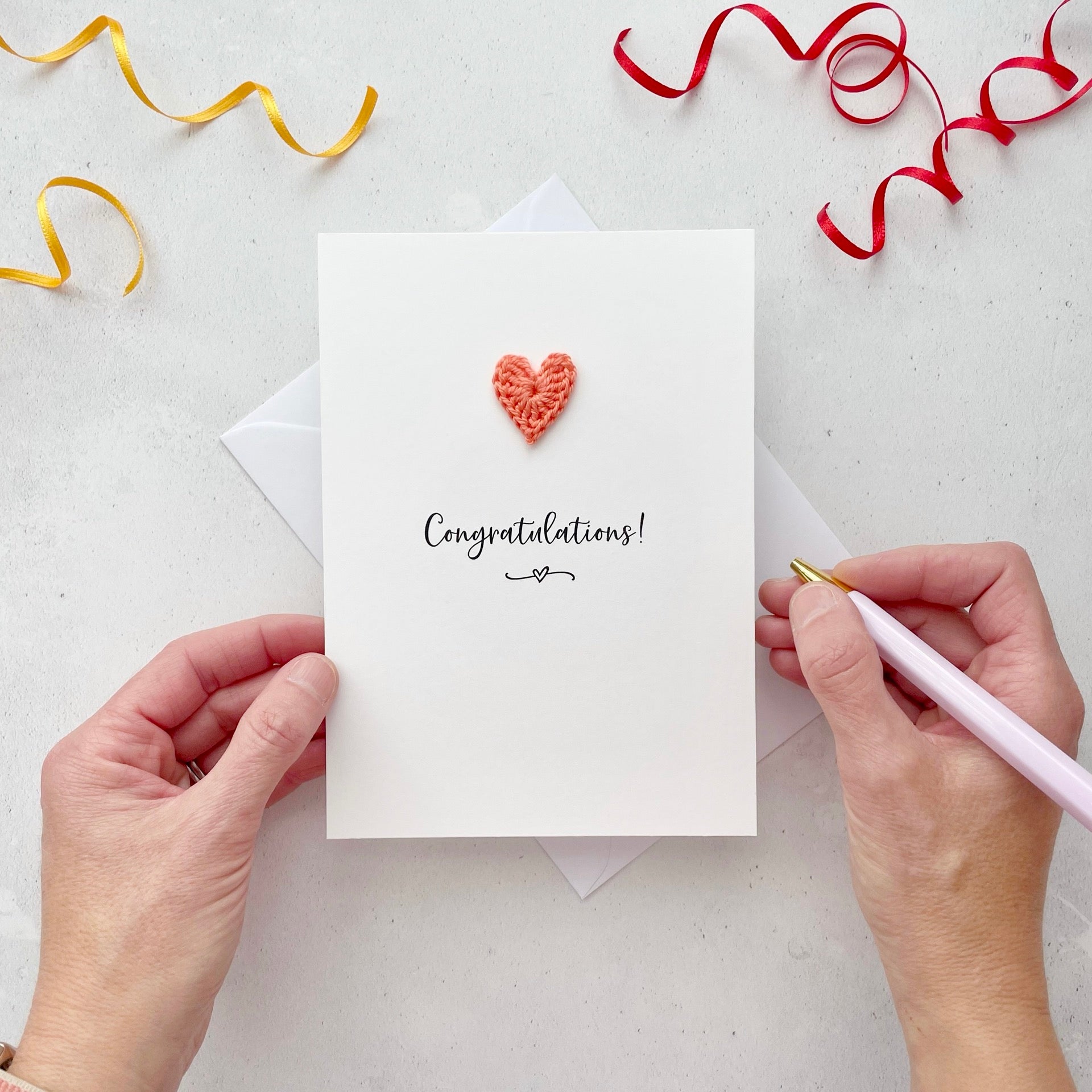 A white portrait-style card featuring the word ‘Congratulations!’ in elegant black cursive at the center. Below the text, a small black squiggle and heart design adds a playful touch. Above the word, a handmade coral coloured crochet heart is carefully attached, providing a pop of colour and texture.