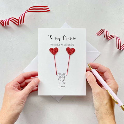 A Christmas card featuring a minimalist design with two figures wearing santa hats holding red glitter heart balloons. The text at the top reads: ‘To my Cousin, with love at Christmas’. The card is placed on a white background with a white envelope partially visible behind it. A hand is seen holding the card, while another hand holds a pink pen. A red and white striped ribbon is positioned in the top corners of the image for decoration.