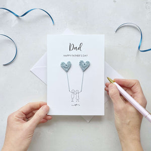 A white Father’s Day card featuring a minimalist design with two line drawn figures holding pale blue crochet heart balloons. The text at the top reads: ‘Dad, Happy Father’s Day. 