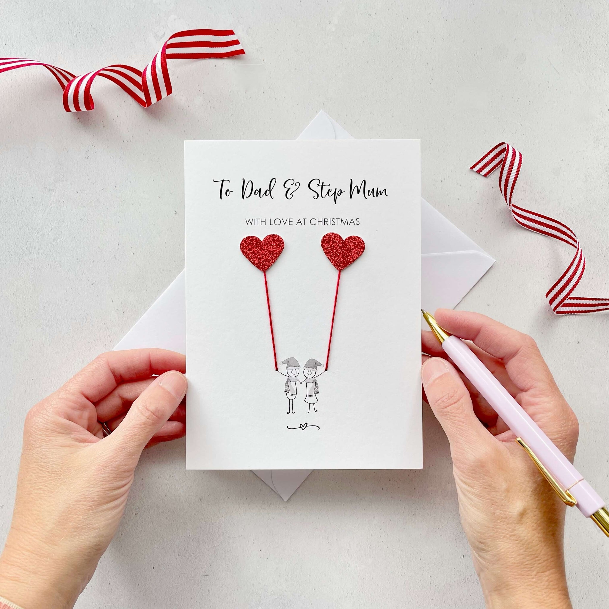 A Christmas card featuring a minimalist design with two figures wearing santa hats holding red glitter heart balloons. The text at the top reads: ‘To Dad & Step Mum, with love at Christmas’. The card is placed on a white background with a white envelope partially visible behind it. A hand is seen holding the card, while another hand holds a pink pen. A red and white striped ribbon is positioned in the top corners of the image for decoration.