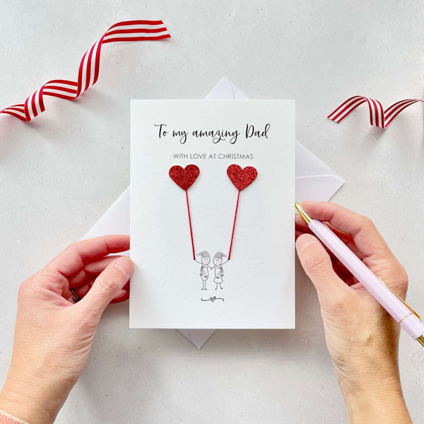 A Christmas card featuring a minimalist design with two figures wearing santa hats holding red glitter heart balloons. The text at the top reads: "To my amazing Dad, With Love at Christmas." The card is placed on a white background with a white envelope partially visible behind it. A hand is seen holding the card, while another hand holds a pink pen. A red and white striped ribbon is positioned in the top corners of the image for decoration.