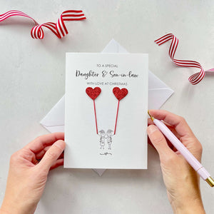 A Christmas card featuring a minimalist design with two figures wearing santa hats holding red glitter heart balloons. The text at the top reads: ‘To a special Daughter & Son-in-law, with love at Christmas’. The card is placed on a white background with a white envelope partially visible behind it. A hand is seen holding the card, while another hand holds a pink pen. A red and white striped ribbon is positioned in the top corners of the image for decoration.