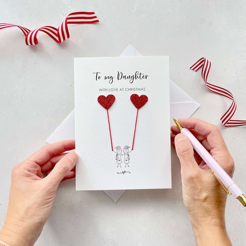 A Christmas card featuring a minimalist design with two figures wearing santa hats holding red glitter heart balloons. The text at the top reads: "To my Daughter, With Love at Christmas." The card is placed on a white background with a white envelope partially visible behind it. A hand is seen holding the card, while another hand holds a pink pen. A red and white striped ribbon is positioned in the top corners of the image for decoration.