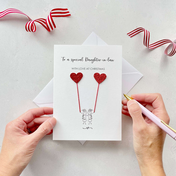 A Christmas card featuring a minimalist design with two figures wearing santa hats holding red glitter heart balloons. The text at the top reads: ‘To a special Daughter in law, with love at Christmas’. The card is placed on a white background with a white envelope partially visible behind it. A hand is seen holding the card, while another hand holds a pink pen. A red and white striped ribbon is positioned in the top corners of the image for decoration.