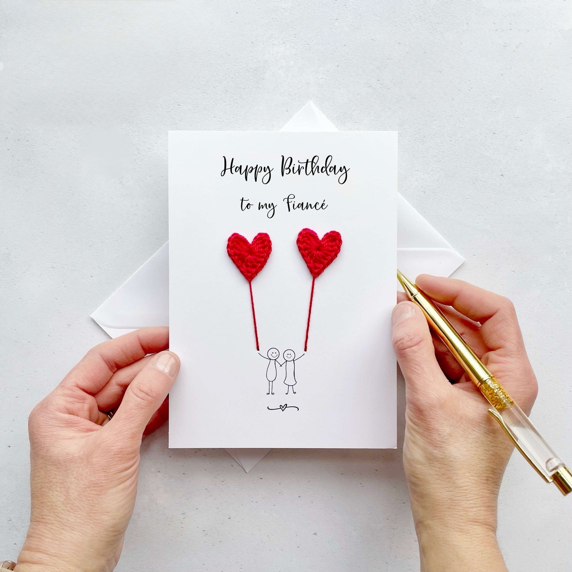 A white birthday card featuring a line drawn couple holding hands and each holding a red crochet heart balloon.  ‘Happy Birthday to my Fiancé’’ is printed at the top of the card in black cursive text.