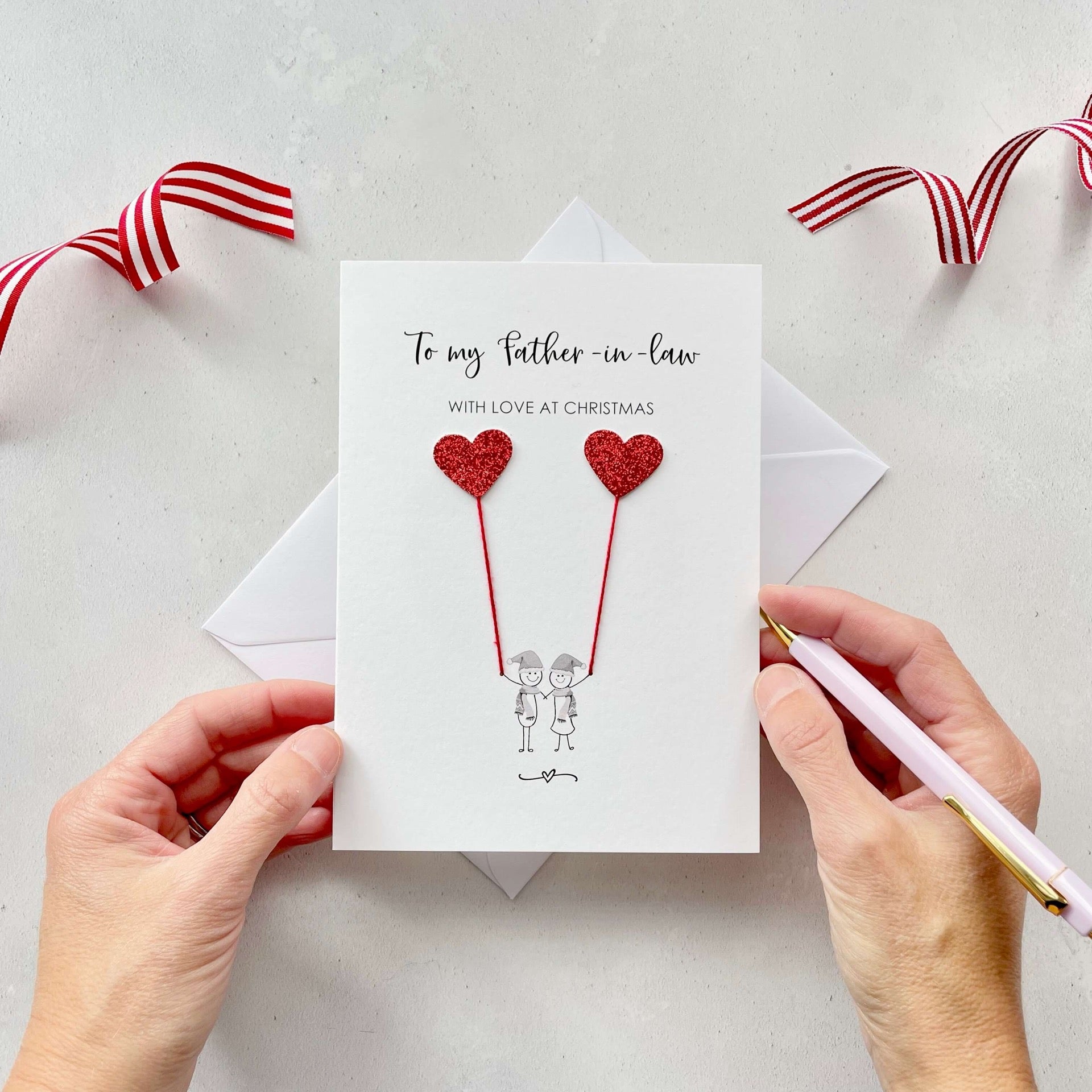 A Christmas card featuring a minimalist design with two figures wearing santa hats holding red glitter heart balloons. The text at the top reads: ‘To my Father in law, with love at Christmas’. The card is placed on a white background with a white envelope partially visible behind it. A hand is seen holding the card, while another hand holds a pink pen. A red and white striped ribbon is positioned in the top corners of the image for decoration.